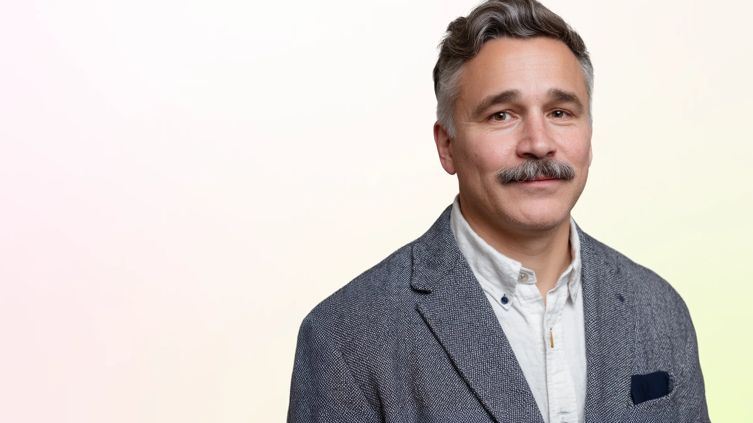Middle-aged man with gray hair and a mustache wearing a gray blazer and white shirt, smiling gently.